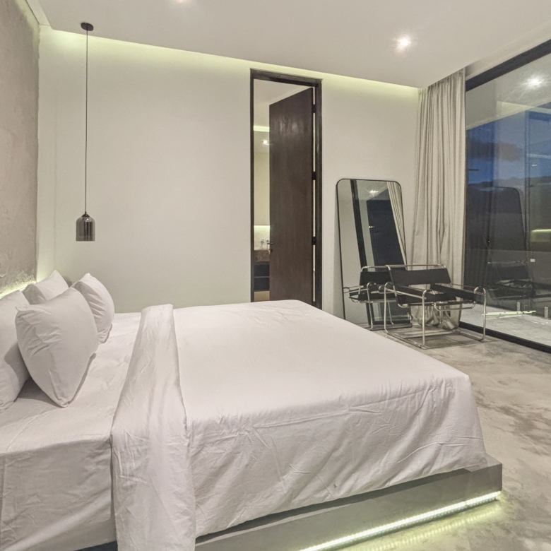 Minimalist hotel bedroom with a white bed, multiple pillows, and a glass wall reflecting the night outside.