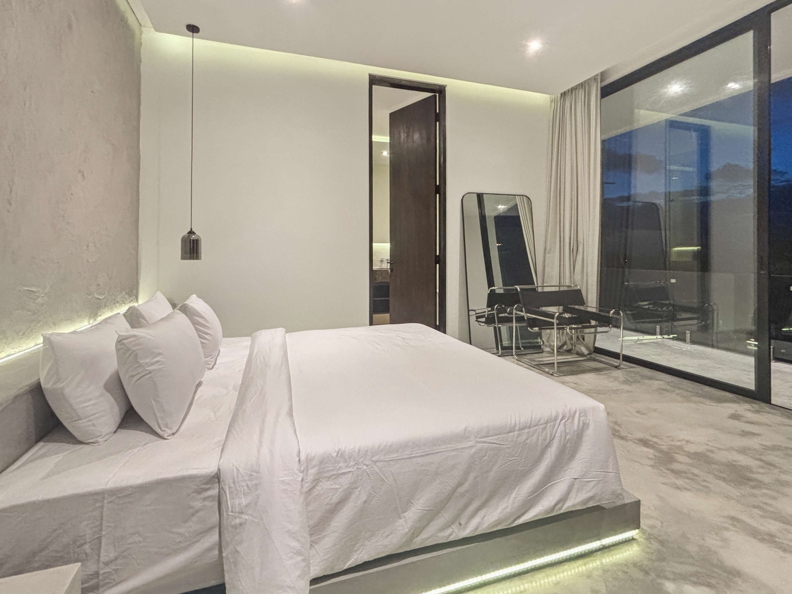 ef0693e2bd1da90bee1dcdcf9f5409ee29a0164c_0938-6a05be07b02ab Minimalist hotel bedroom with a white bed, multiple pillows, and a glass wall reflecting the night outside.
