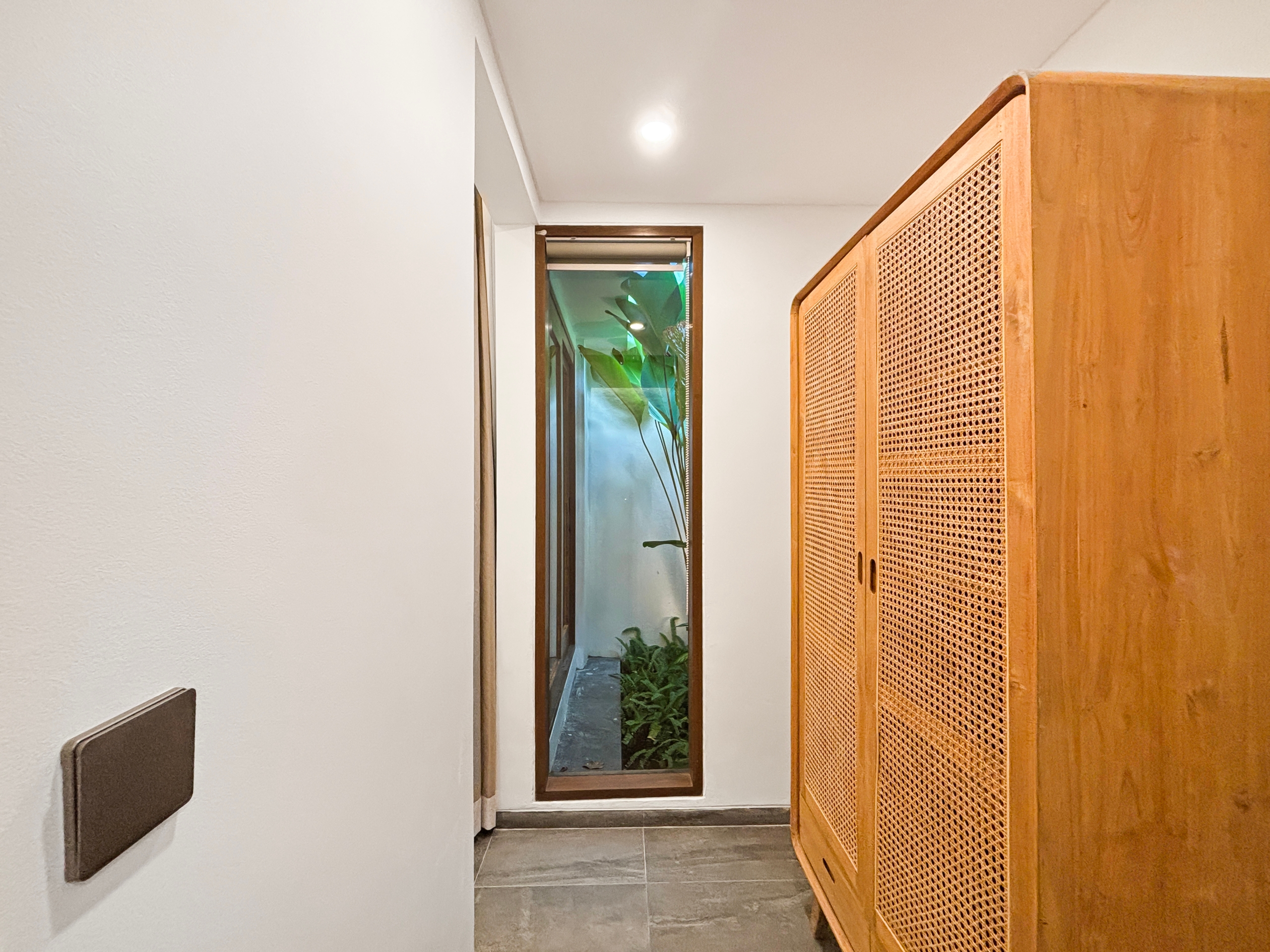 efb60556f7353c53673ebd2ee7c631c748b3bf24 Narrow indoor hallway with white walls, tile floor, ceiling light, and a tall wooden cabinet with woven cane panels on the right.