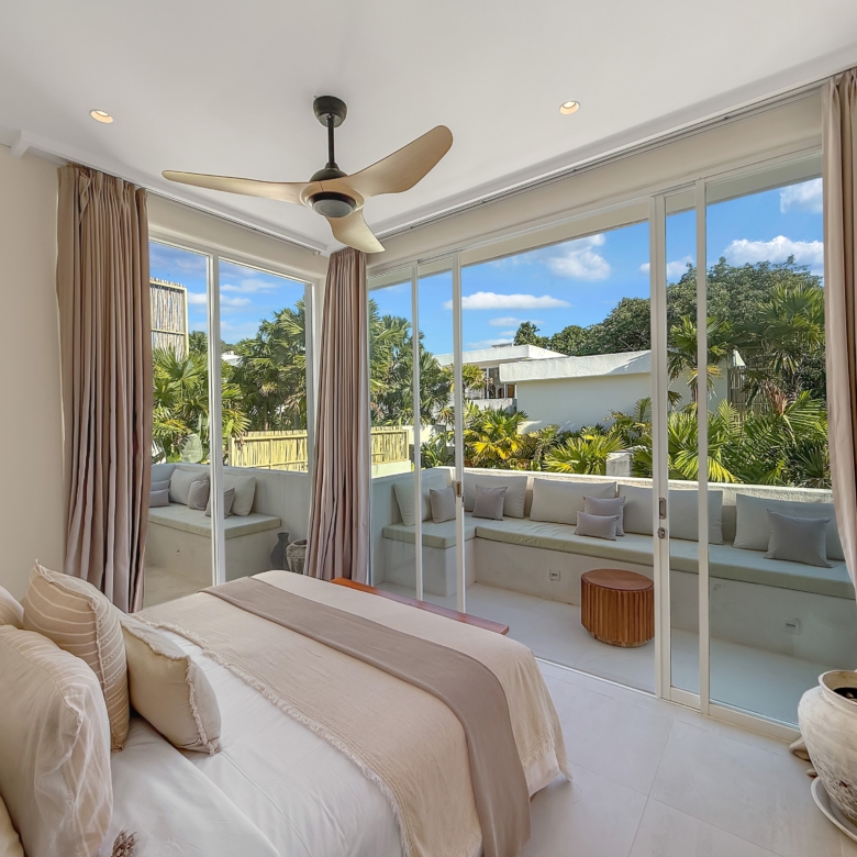 Bright modern bedroom with beige bedding and floor-to-ceiling glass doors opening to a patio with palm trees outside.