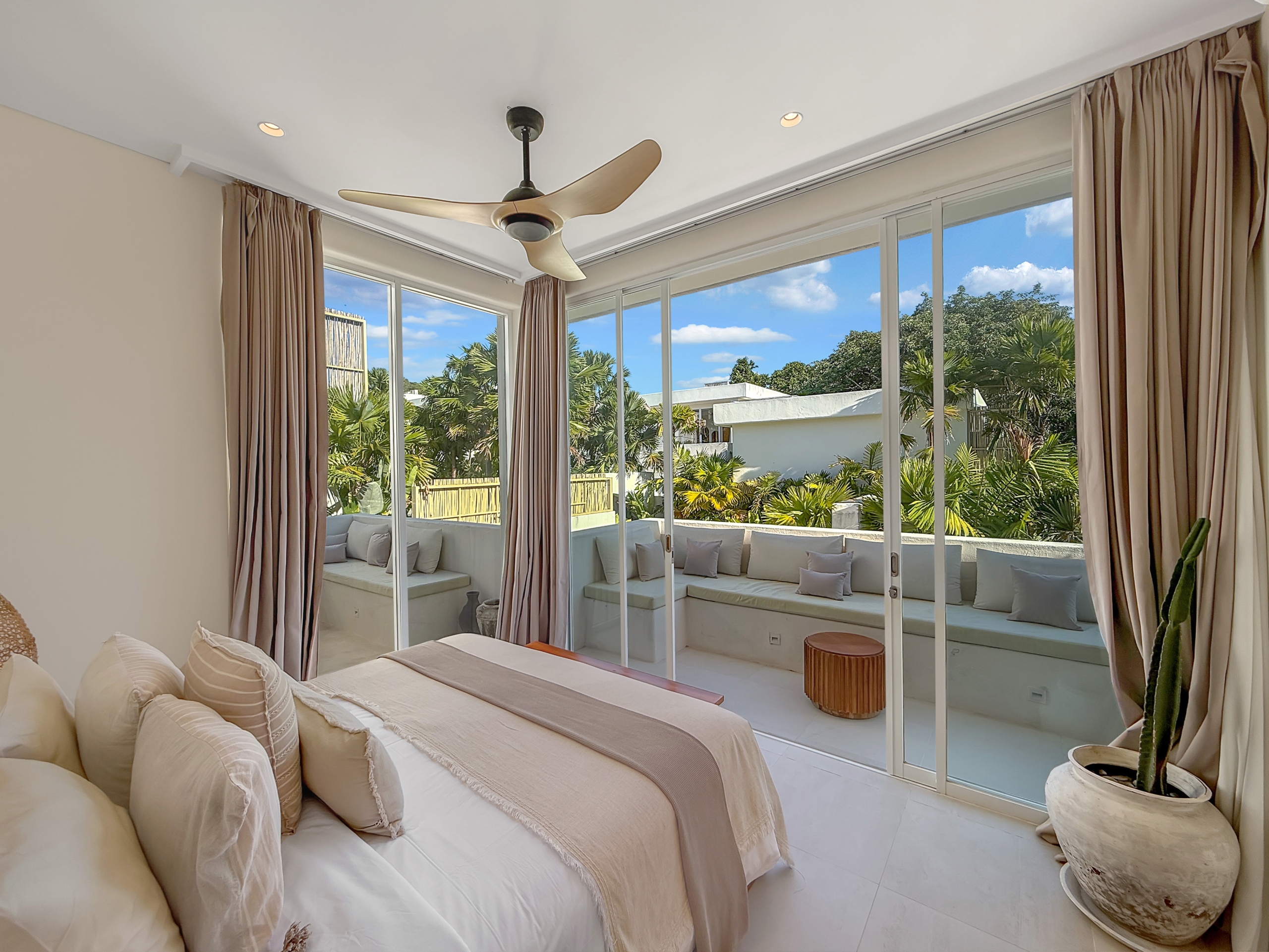 f2a249193917d2ca14e63b7f3d92d53b00977577 Bright modern bedroom with beige bedding and floor-to-ceiling glass doors opening to a patio with palm trees outside.