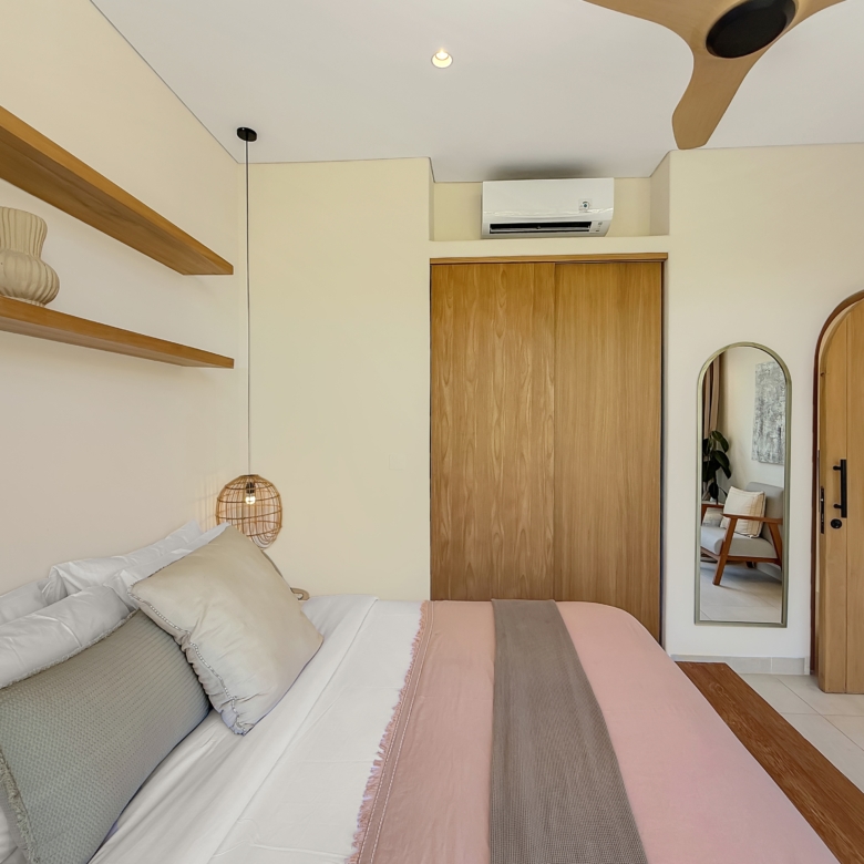 Hotel-style bedroom with a bed, pillows, and pastel pink bed runner; wooden shelves and decor to the left, and a wooden door with a full-length mirror on the right.