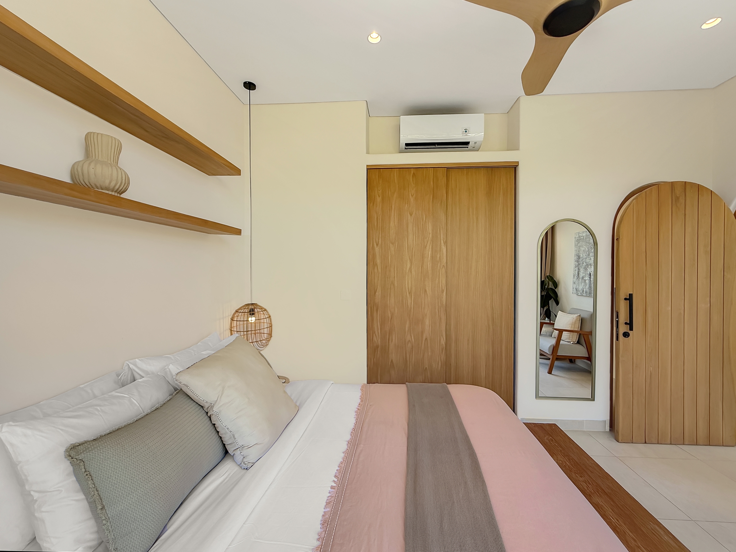 f4aec89c13d8f89565527a3c98c38a3d8cfb9383 Hotel-style bedroom with a bed, pillows, and pastel pink bed runner; wooden shelves and decor to the left, and a wooden door with a full-length mirror on the right.