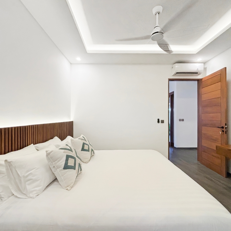Hotel bedroom with white bedding and a dark wood headboard, ceiling fan, and doorway to the right; a wall-mounted TV visible.