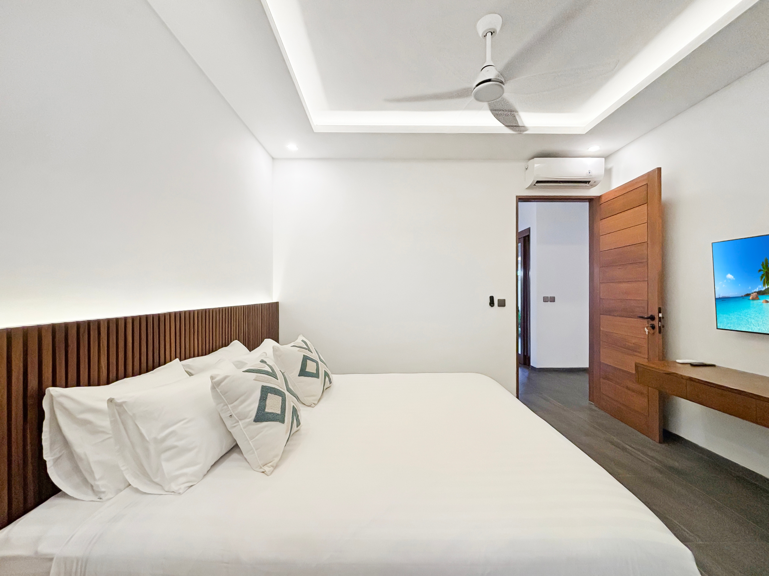 f6301310a3e14a3cbb8adc991cdf13aa7088bdc2 Hotel bedroom with white bedding and a dark wood headboard, ceiling fan, and doorway to the right; a wall-mounted TV visible.