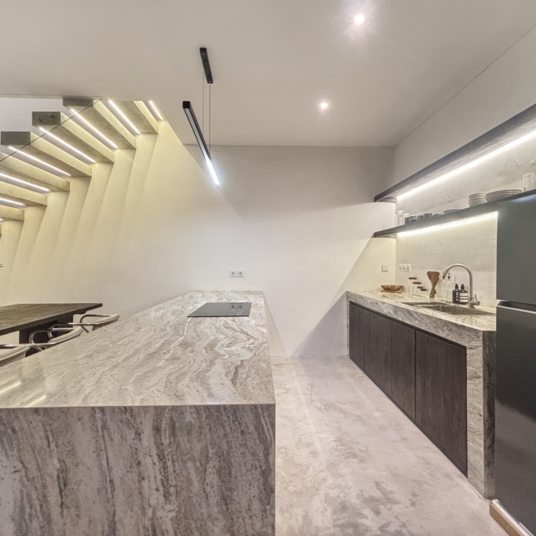 Modern kitchen with a large marble island, dark base cabinets, and a sleek staircase along the left wall; bright, minimalist design.