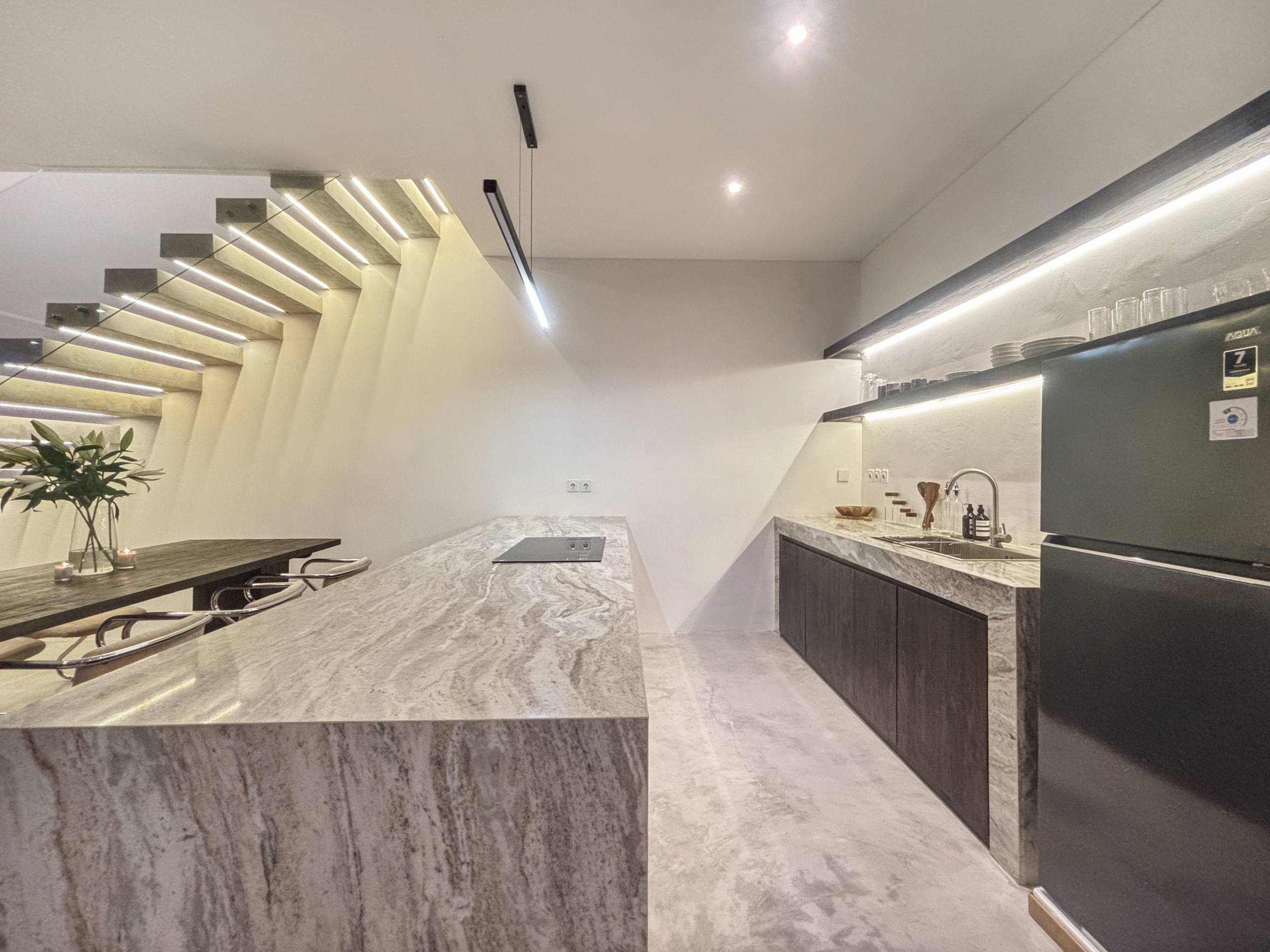 f77c3dacf9f2ab45309989aa7b6ecaa442058fd7_1006-6a05be068a640 Modern kitchen with a large marble island, dark base cabinets, and a sleek staircase along the left wall; bright, minimalist design.
