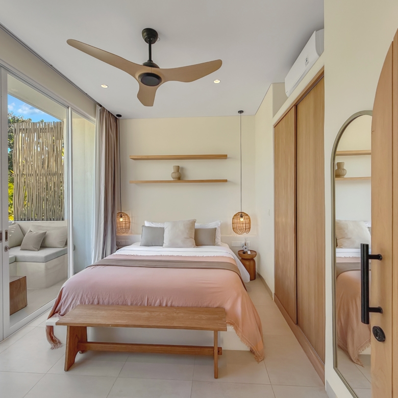 Bright bedroom with blush-pink duvet, wooden bench at the foot, and two hanging pendant lamps flanking a white bed against a wall with wood shelves and a mirror-door closet.