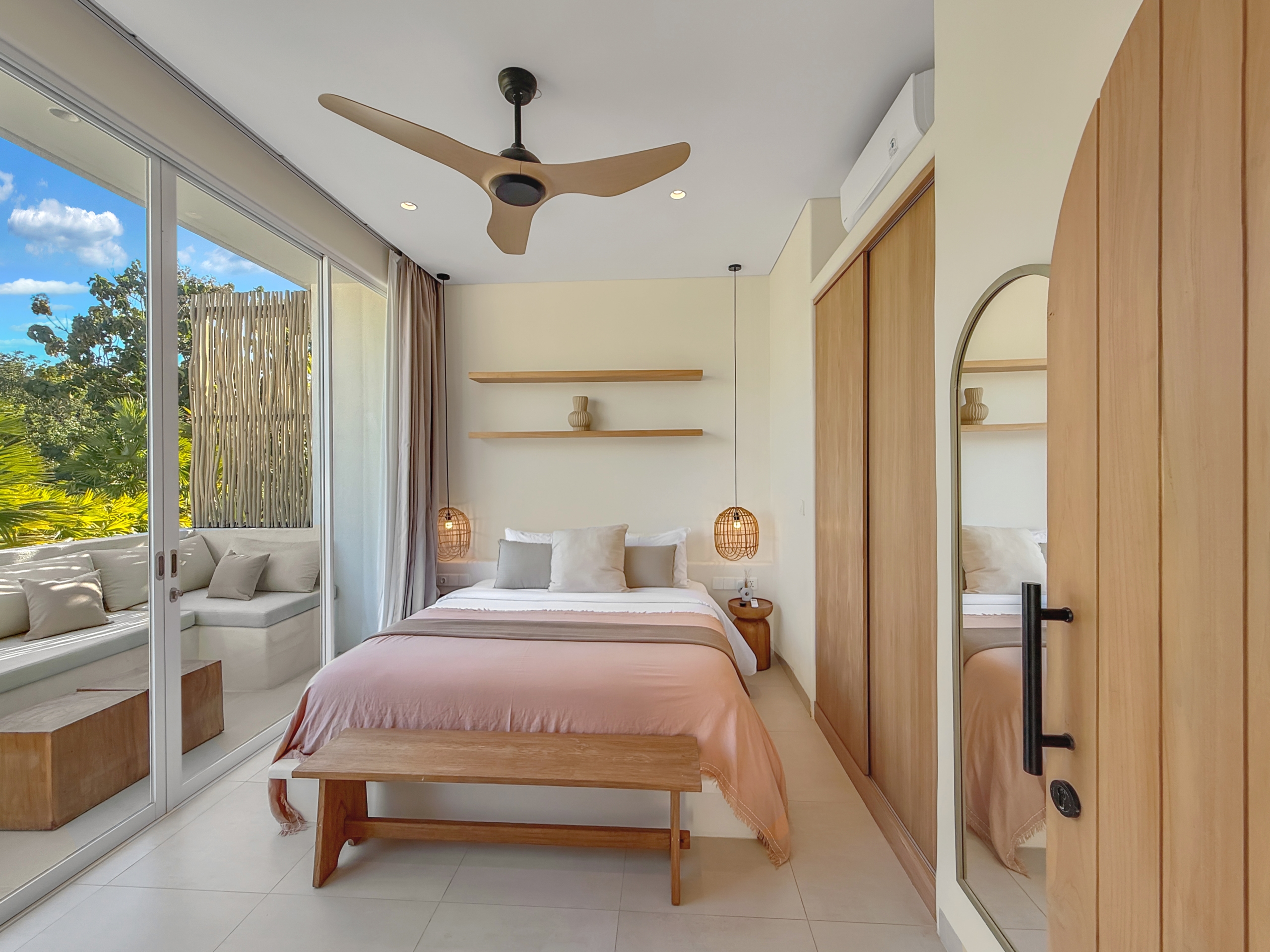 f78b3cc6e04bb183e2519e288714cd83d441794b Bright bedroom with blush-pink duvet, wooden bench at the foot, and two hanging pendant lamps flanking a white bed against a wall with wood shelves and a mirror-door closet.