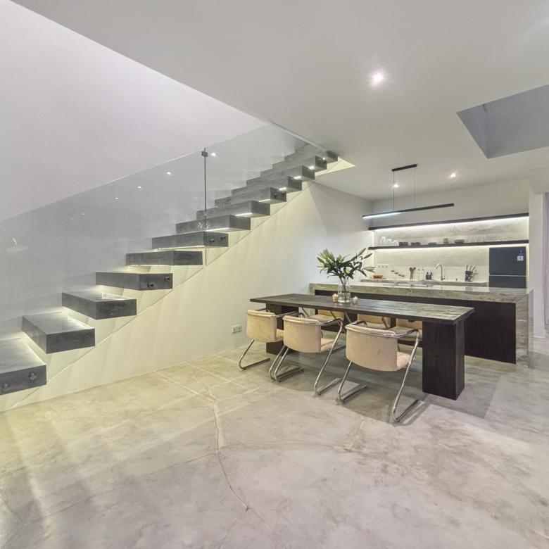 Modern open-plan kitchen and dining area with a floating staircase and glass railing, marble floors, and a minimalist table and chairs in the foreground.