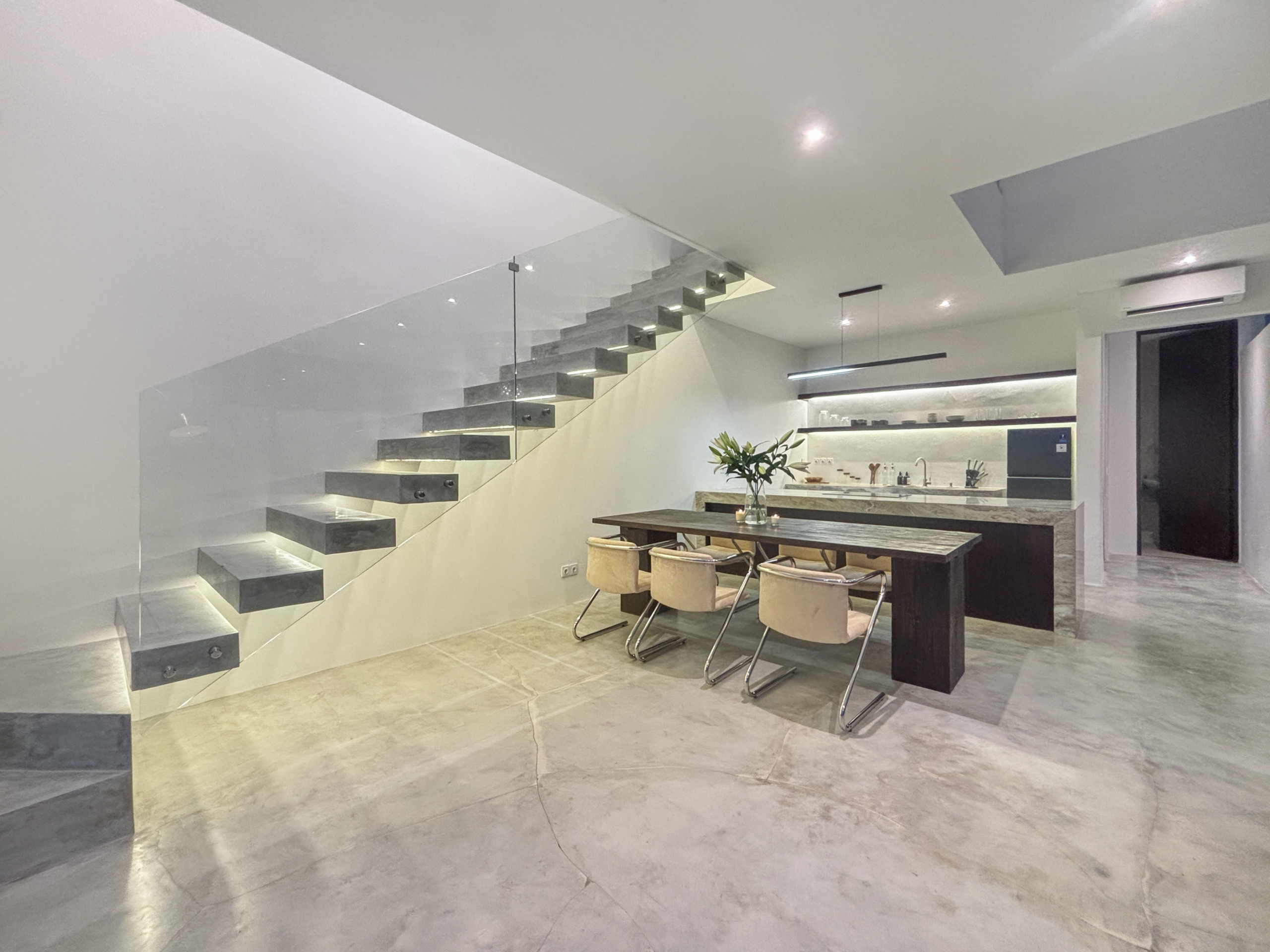 f78d0d5c5656694abbad3c739c9e5ef23c8298f2_0998-6a05be063627d Modern open-plan kitchen and dining area with a floating staircase and glass railing, marble floors, and a minimalist table and chairs in the foreground.