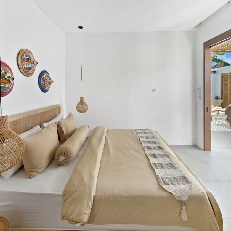 Beige bed with multiple pillows and woven pendant lights in a bright white bedroom, with an open patio door visible to the outside.