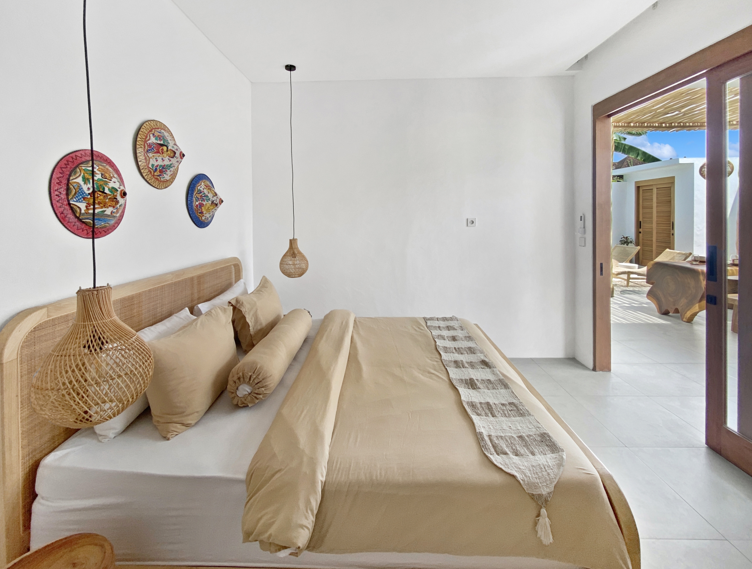 f97e9d5366ef11c6f907615561d628fcbf6a9408_2289-68d5cfaeb3f2d Beige bed with multiple pillows and woven pendant lights in a bright white bedroom, with an open patio door visible to the outside.
