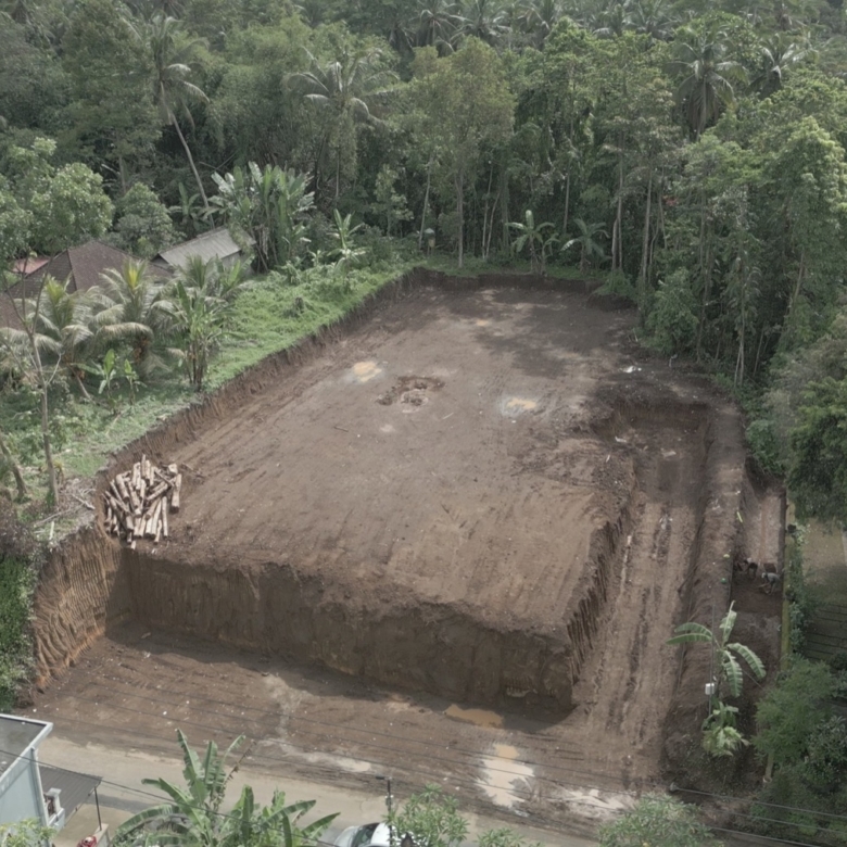 Aerial view of a construction site in a tropical area, with a large dirt lot surrounded by trees and nearby houses on the left.