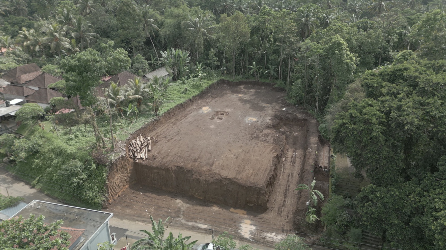0073_Actual_Drone_Frame_Date_Unverified Aerial view of a construction site in a tropical area, with a large dirt lot surrounded by trees and nearby houses on the left.