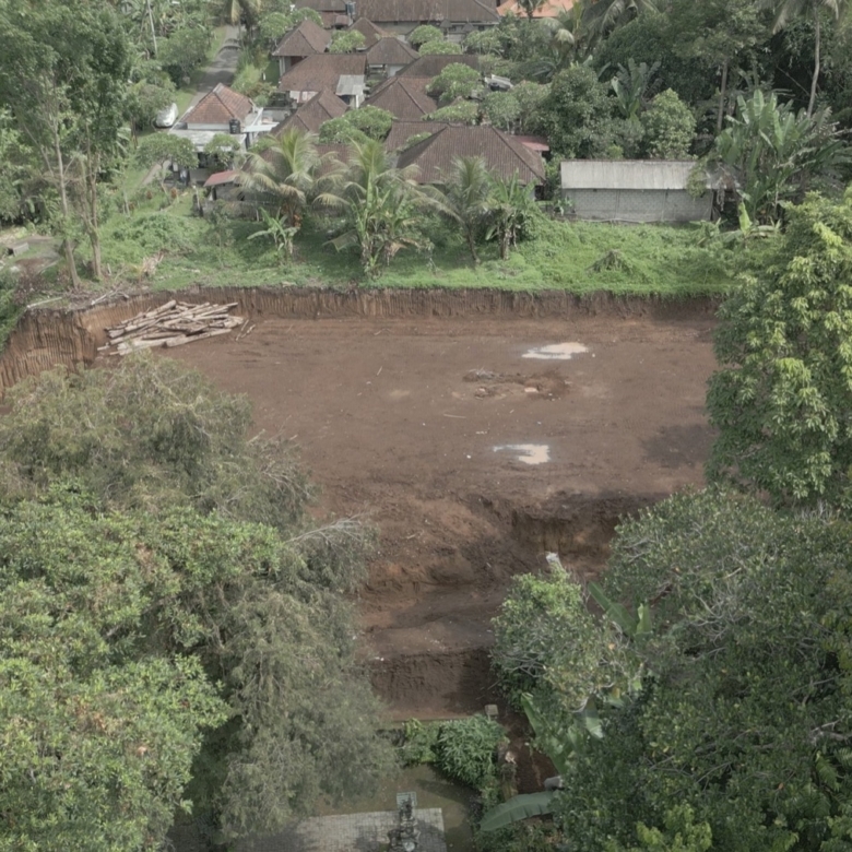 A cleared brown construction site surrounded by dense trees, with a pile of logs to the left and a village of thatched-roof houses beyond.