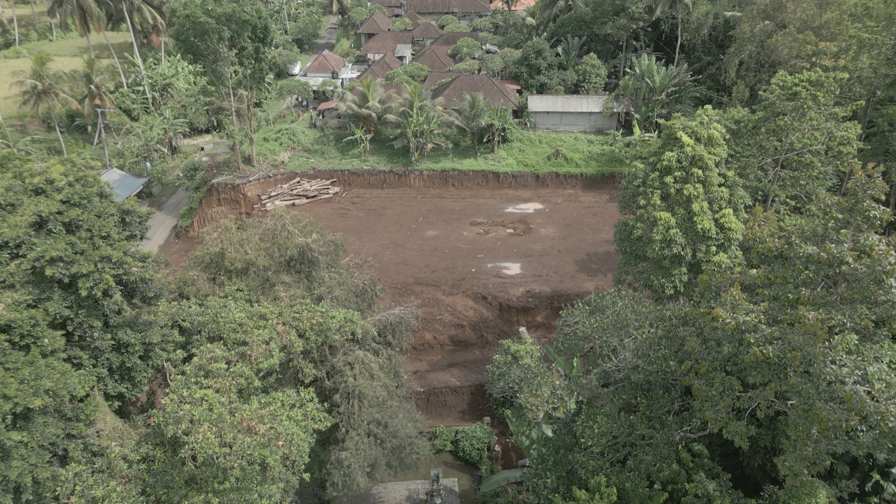 0075_Actual_Drone_Frame_Date_Unverified A cleared brown construction site surrounded by dense trees, with a pile of logs to the left and a village of thatched-roof houses beyond.