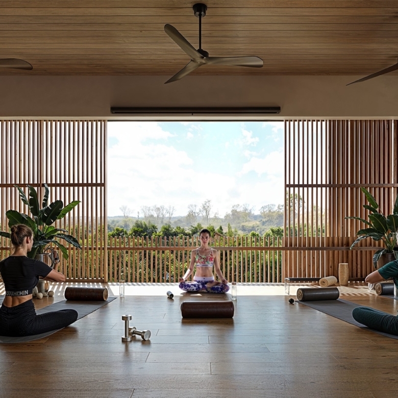 Yoga class in an open, sunlit studio with wooden slats; participants sit on mats in lotus pose around a central instructor in a colorful outfit, plants nearby.